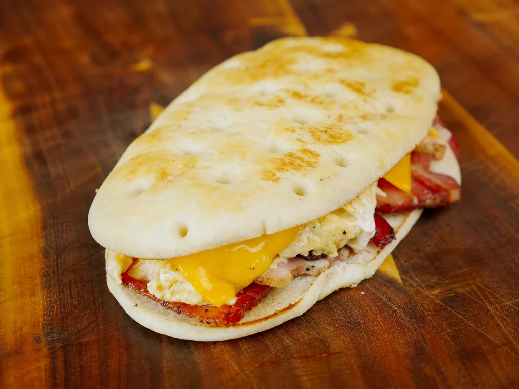 Bacon & Egg Sandwich – Fladenbrot mit Rührei, Bacon und geschmolzenem Cheddar, angeschnitten auf Holzbrett