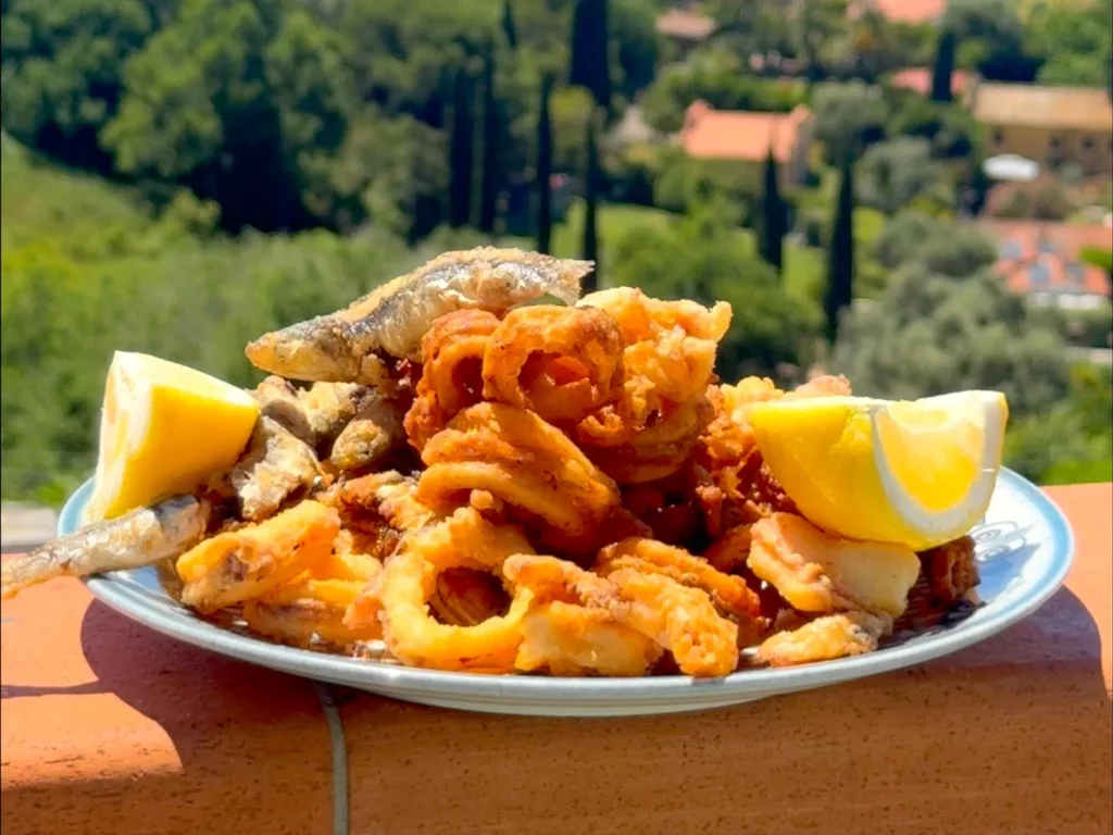 Calamari Fritti – knusprig frittierte Calamari-Ringe mit Zitronenspalten auf Teller, im Hintergrund mediterrane Küstenlandschaft