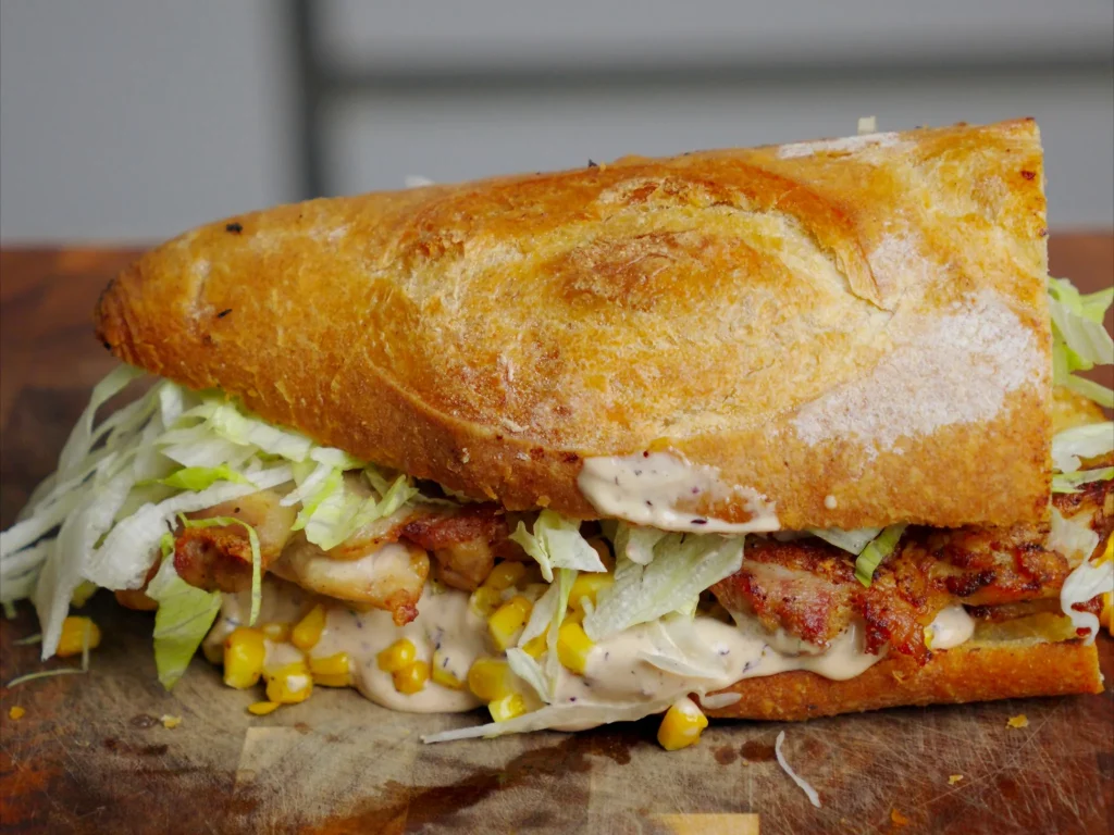 Hähnchen Sandwich – Baguette mit Hähnchen, Salat, Mais und Aioli, angeschnitten auf Holzbrett