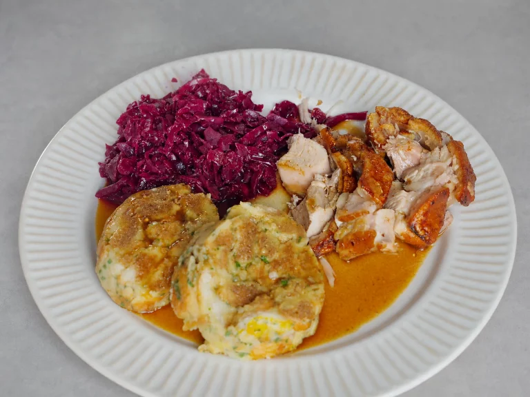 Krustenbraten mit Dunkelbiersoße und Serviettenknödel – knusprige Schweinekruste mit Serviettenknödeln, Rotkohl und Soße auf weißem Teller