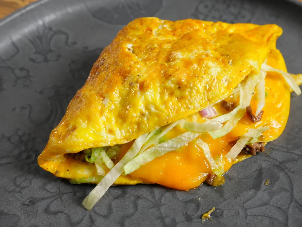 Omelette-Wrap Big Mac Edition mit Hackfleisch, Cheddar, Salat und roten Zwiebeln, aufgeschnitten auf dunklem Teller