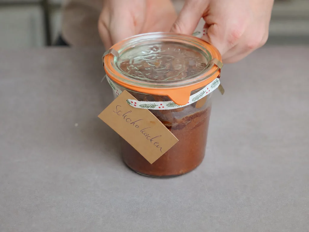 Schokoladenkuchen im Glas mit weihnachtlichem Deckel, Hand hält das Glas mit Anhänger "Schokokuchen"
