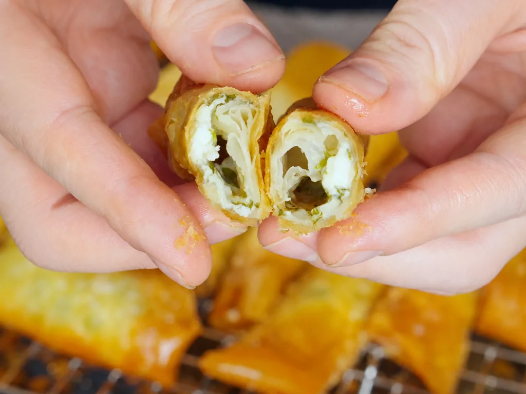 Sigara Börek mit Feta-Füllung, aufgebrochen in der Hand gehalten, im Hintergrund weitere Röllchen
