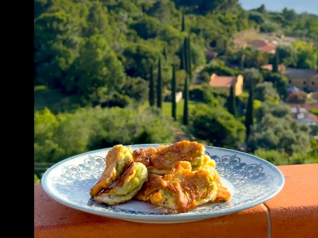 Knusprig ausgebackene Zucchiniblüten auf gemustertem Teller, im Hintergrund toskanische Landschaft