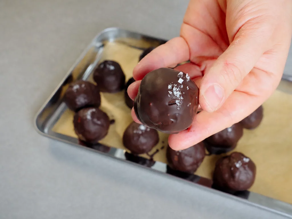 Snickers Energy Balls – Schokoladen-Energy-Balls mit Salzflocken auf einem Blech, eine Kugel in der Hand