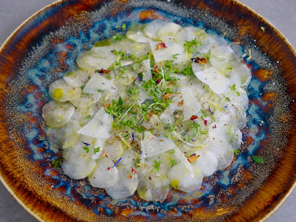 Spargel Carpaccio aus dünn gehobeltem weißem Spargel mit Kresse, essbaren Blüten und Käsespänen auf gesprenkeltem Keramikteller