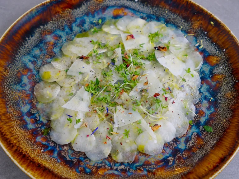 Spargel Carpaccio aus dünn gehobeltem weißem Spargel mit Kresse, essbaren Blüten und Käsespänen auf gesprenkeltem Keramikteller