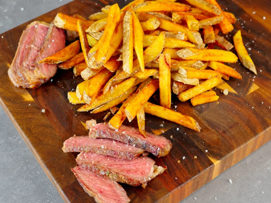 In Scheiben geschnittenes Steak mit knusprigen Fries auf Holzbrett, mit Fleur de Sel bestreut