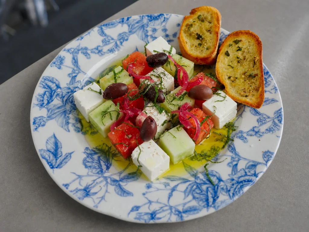 Wassermelonen-Feta-Salat mit Gurke, roten Zwiebeln, Oliven und Minze, dazu geröstetes Knoblauch-Baguette auf blau-weißem Blumenteller